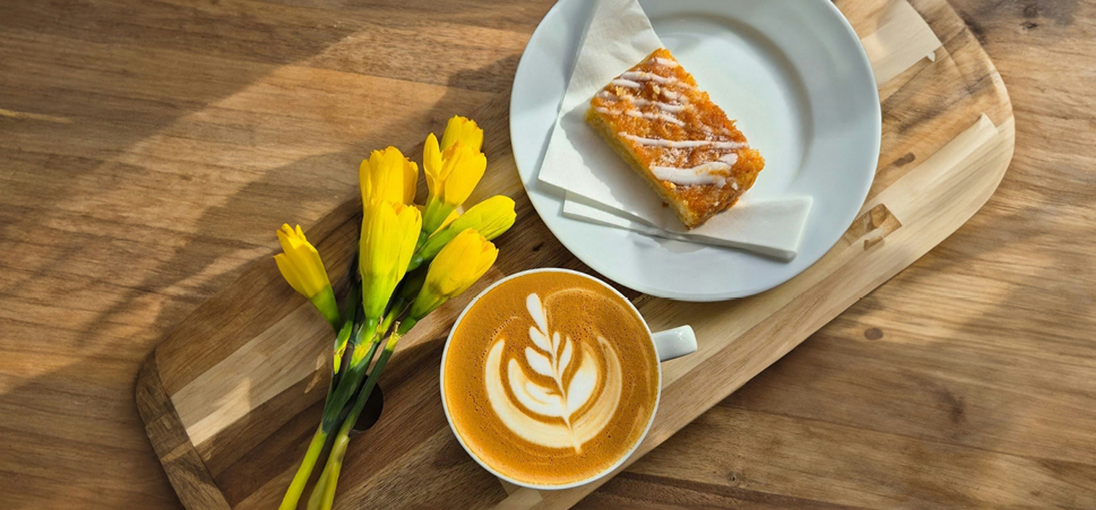 A cup of coffee with a leaf design in foam on a wooden table, alongside a piece of cake and yellow tulips.