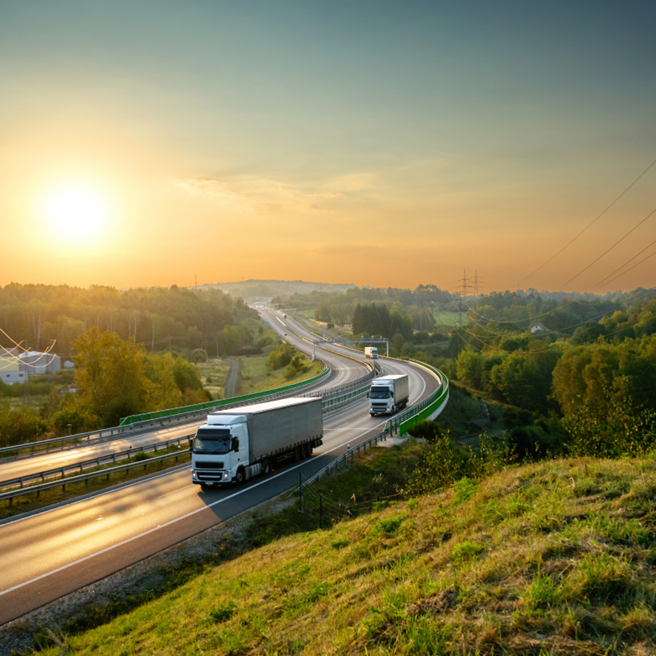 Early morning highway scene with two white trucks driving, a sunrise background with greenery, and power lines in the distance.