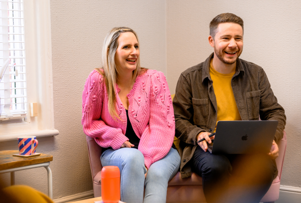 Two colleagues in a casual office setting, a woman in a pink cardigan and a man with a laptop, laughing and enjoying a conversation.