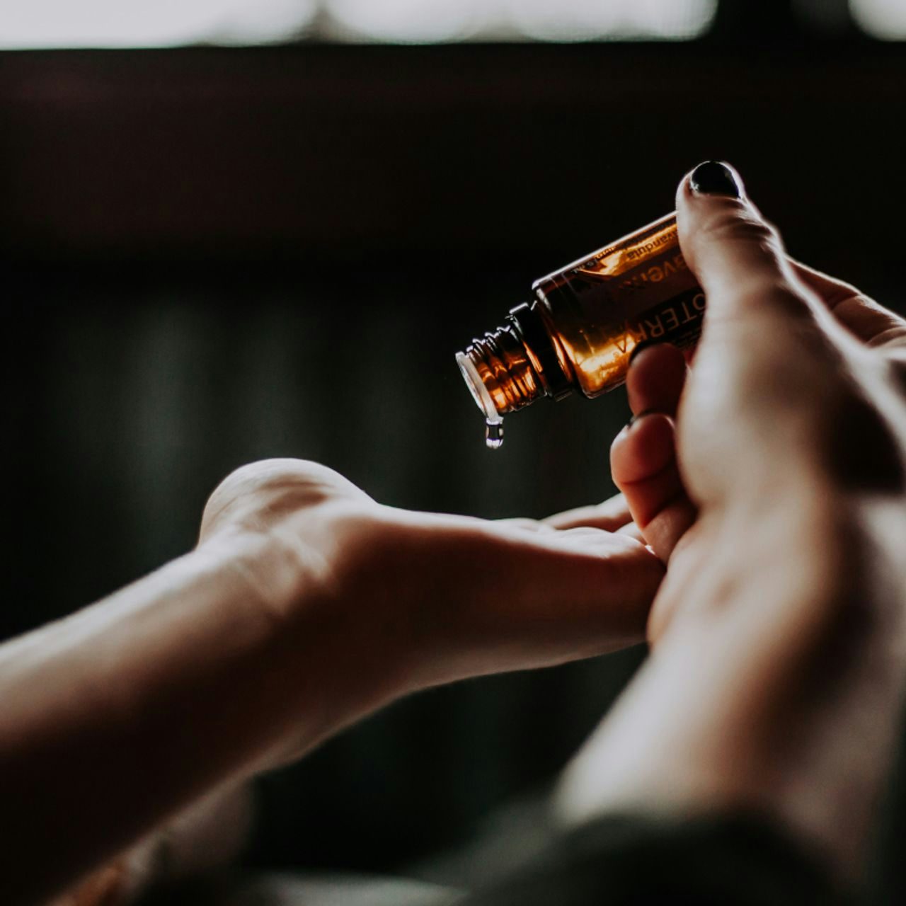 Close-up of hands using a dropper to apply essential oil from an amber bottle, with a focus on a single drop, in a serene setting suggestive of wellness and self-care.