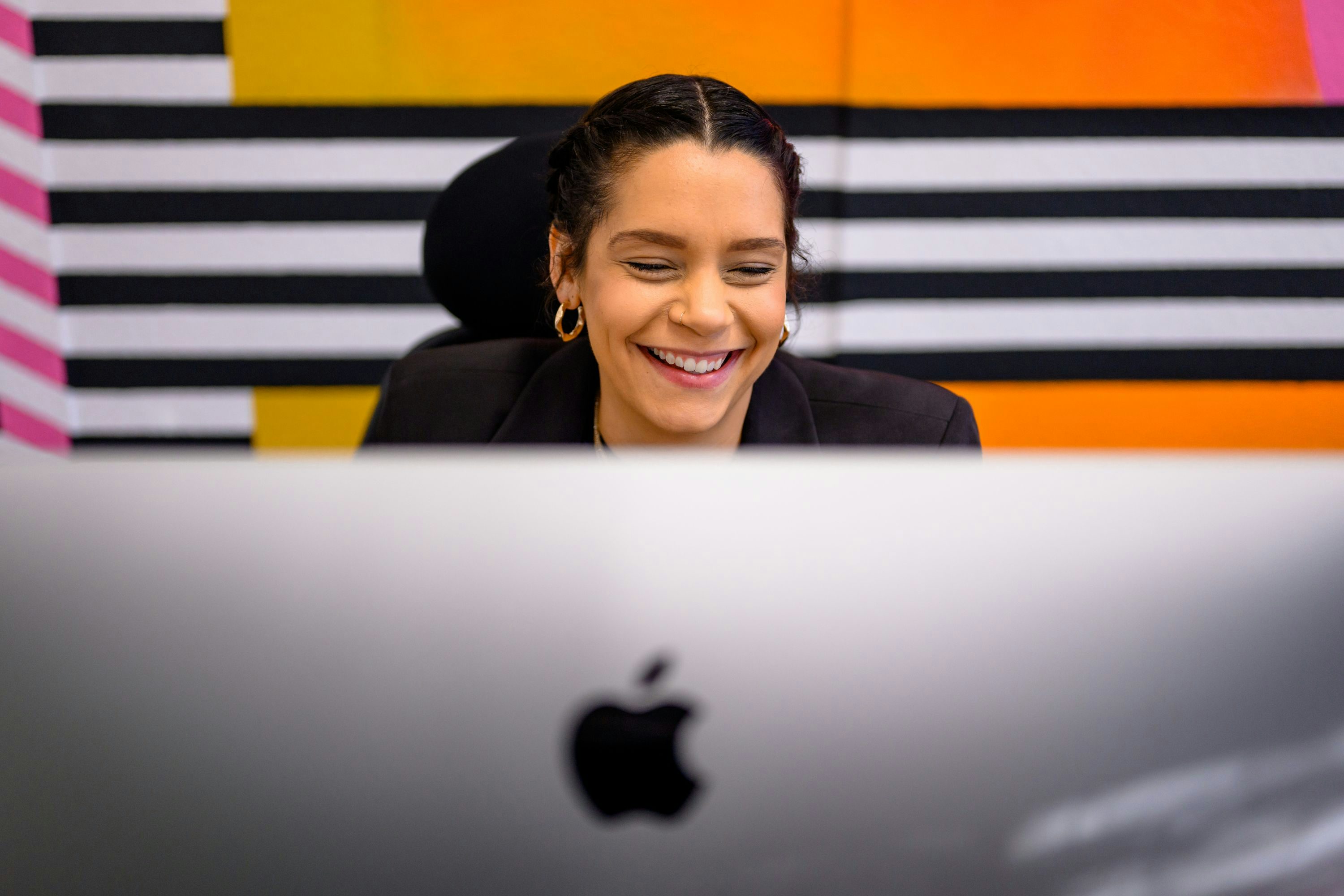 A professional woman wearing a black blazer is smiling and working at her desk. The foreground shows the back of an Apple computer monitor, and the background features a colourful striped wall with yellow, orange, black and white stripes.